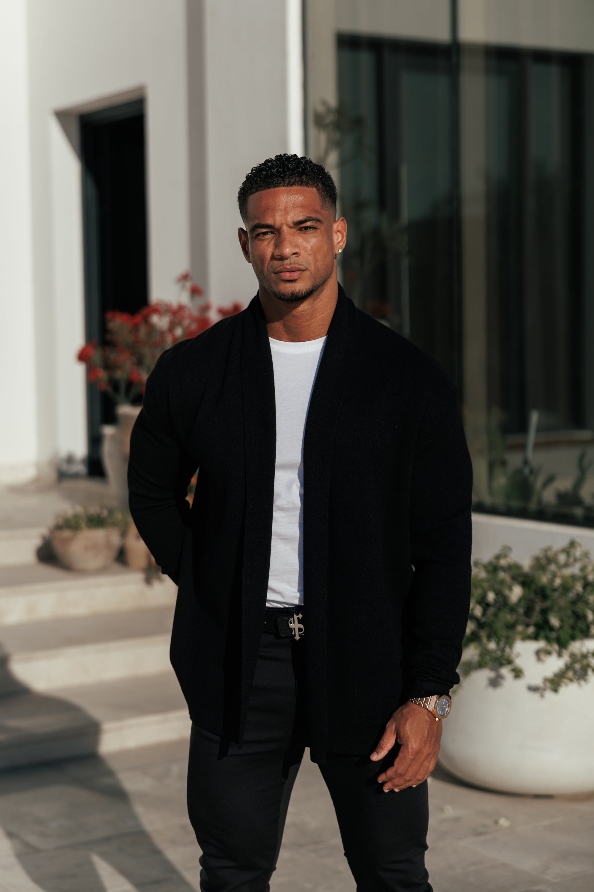 Man in a black jacket standing outdoors with plants and a building in the background