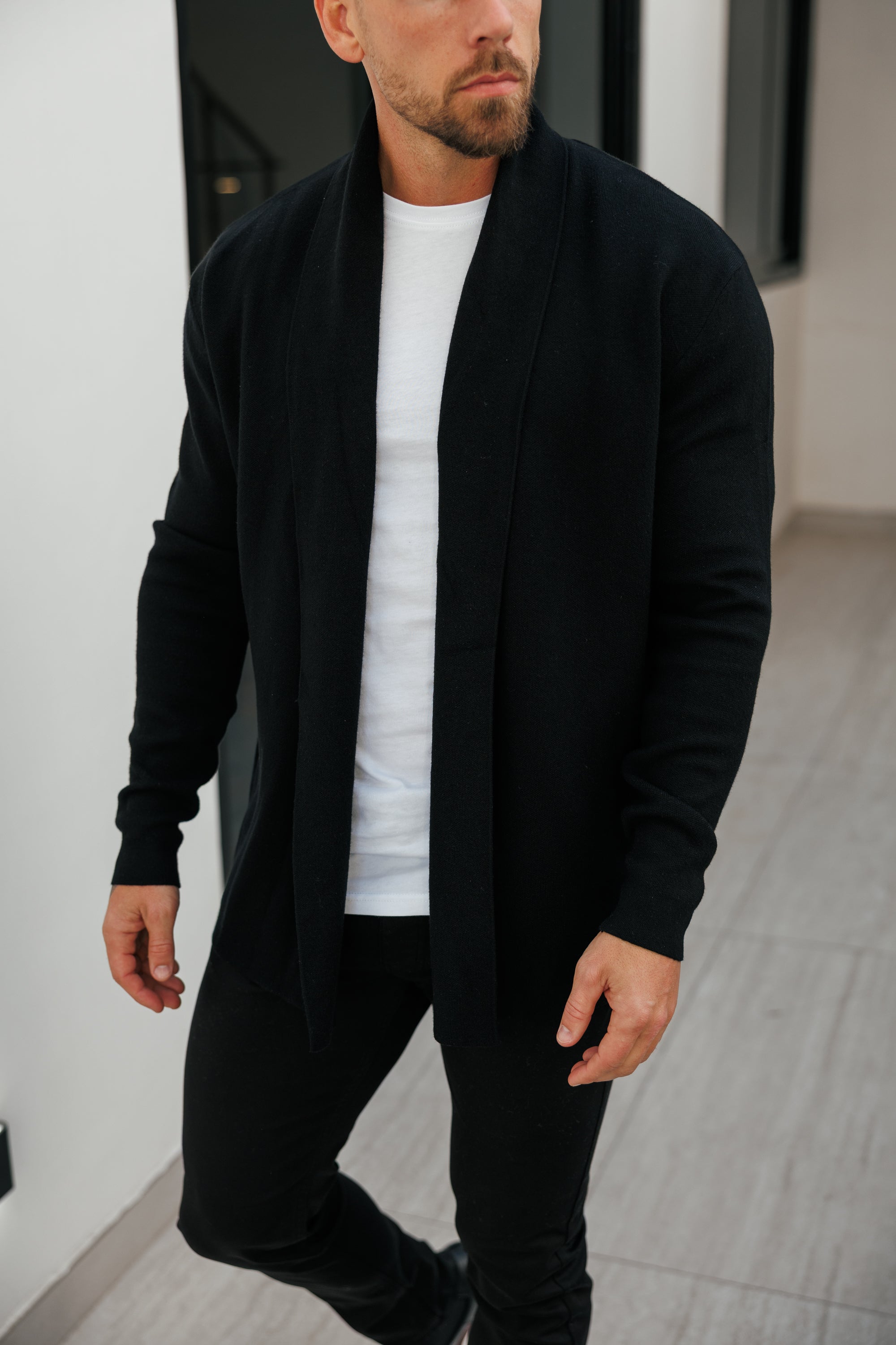 Man wearing a black cardigan over a white shirt and black pants in an urban setting.