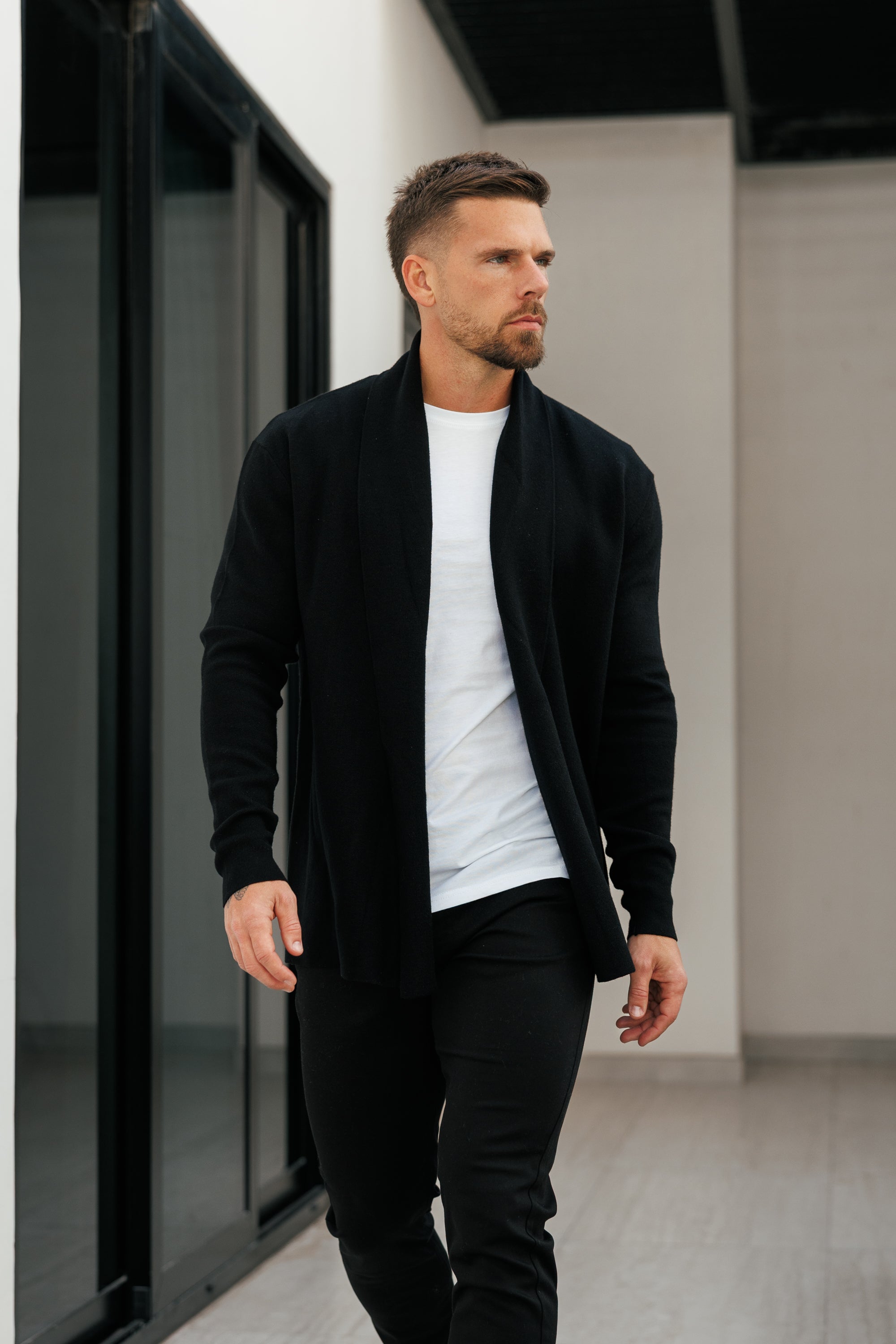 Man wearing a black cardigan over a white shirt and black pants in an indoor setting.