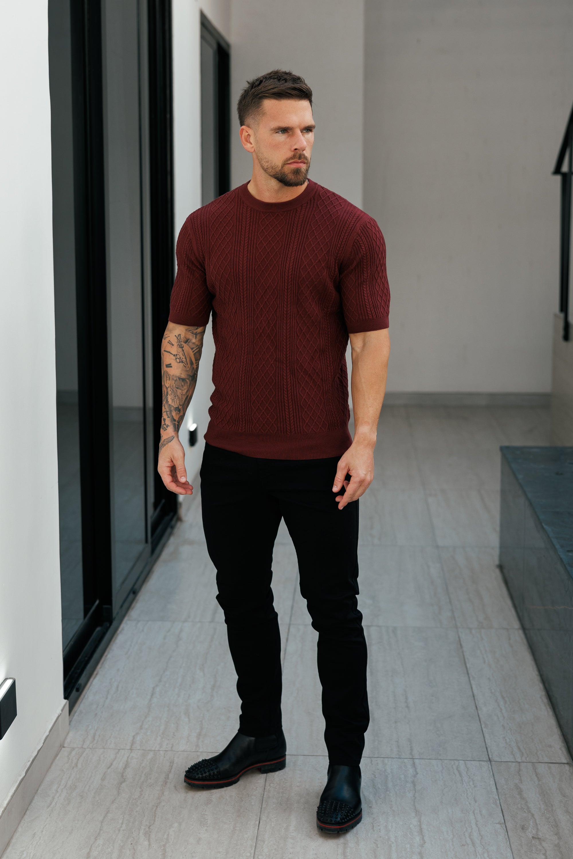Man wearing a burgundy sweater and black pants standing in a modern indoor setting.